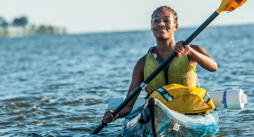 A young person smiles confidently as they paddle a kayak on very blue water.